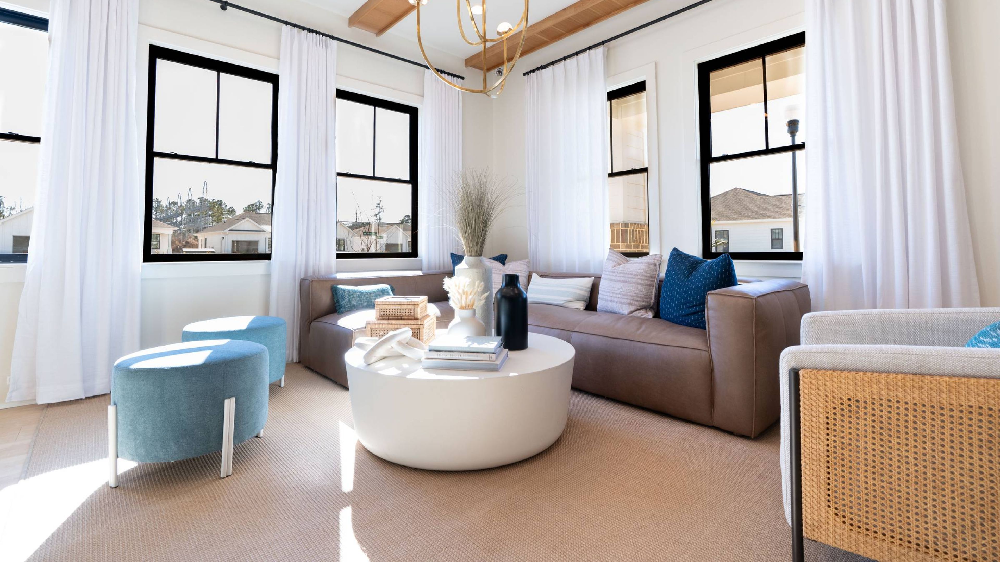 Hawthorne Cottages at Leland bright and airy living room featuring a cozy sofa, stylish ottomans, and large windows with sheer curtains.